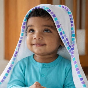 May include: A smiling baby wearing a light blue long-sleeved shirt and a white hooded towel. The towel is trimmed with a pattern of blue and purple squares. The background is a warm, neutral color.