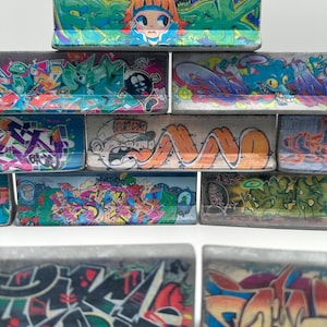 Könnte beinhalten: Ein Set aus Miniatur-Beton-Skate-Rampen mit bunten Graffiti-Designs. Die Rampen sind übereinander gestapelt und bilden einen kleinen Skatepark. Die Graffiti-Designs umfassen abstrakte Muster, Comicfiguren und Wörter.