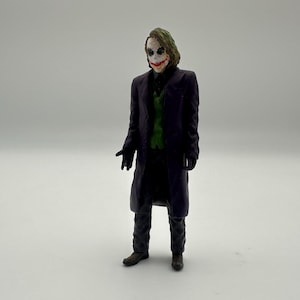May include: A plastic action figure of the Joker from the Batman movie, wearing a purple coat, a green vest, and a white mask with a red smile.