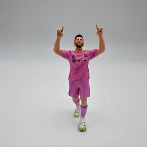 Pode incluir: Estatueta de um jogador de futebol com uniforme rosa com o texto "XB TO" no peito. O jogador está com os dois braços levantados, com os dedos indicadores apontando para cima. A figura usa meias brancas, meias listradas rosa e preto e sapatos brancos e verdes.