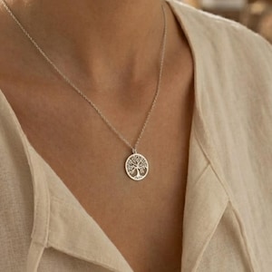 May include: A silver necklace featuring a circular pendant with a tree of life design. The pendant hangs from a delicate chain. The necklace is worn against a light-colored top.