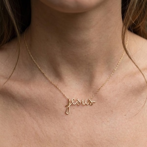 Peut inclure: Un collier doré avec le mot "Jesus" écrit en cursive. Le pendentif est attaché à une chaîne délicate. Le collier est porté autour du cou.