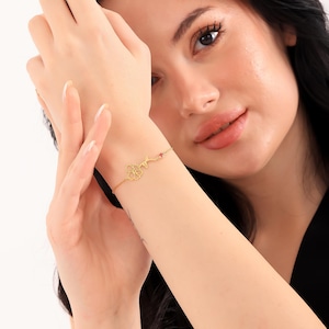 Peut inclure: Un bracelet en or avec un délicat charme de fleur et une petite pierre précieuse rouge. Le bracelet est porté au poignet d'une femme.