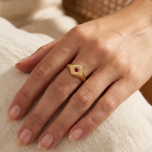May include: A gold ring with a square face and a small, round, red gemstone. The ring is on a hand with well-manicured nails. The ring's design includes a starburst pattern around the gemstone.