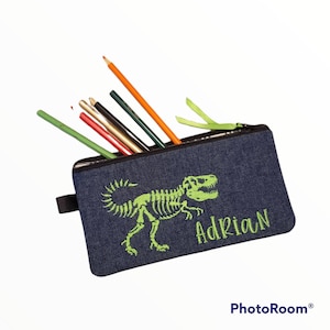 May include: A blue denim pencil case with a green dinosaur skeleton design and the name "Adrian" embroidered on it. The case is filled with colored pencils.