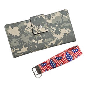 May include: A rectangular wallet featuring a gray and beige digital camouflage pattern with a snap-button closure, accompanied by a wristlet keychain strap displaying a red, white, and blue American flag-inspired pattern with a metal ring.
