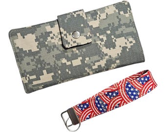 May include: A rectangular wallet featuring a gray and beige digital camouflage pattern with a snap-button closure, accompanied by a wristlet keychain strap displaying a red, white, and blue American flag-inspired pattern with a metal ring.