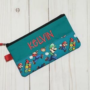 May include: Teal pencil case with a black zipper and red pull tab. The top section has the name "KELVIN" embroidered in red. The bottom section features a Super Mario Bros. print with Mario and Luigi.