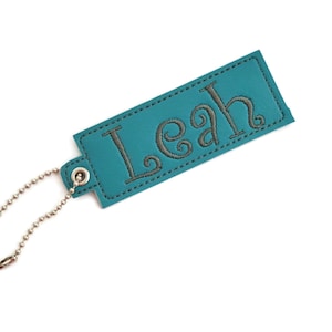 May include: Teal rectangular luggage tag with the name "Leah" embroidered in gray thread. The tag has a silver ball chain attached for easy fastening. The tag is made of a smooth material and has a stitched border.