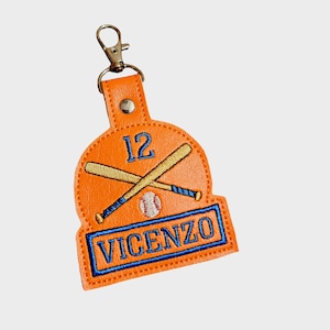 May include: Orange baseball-themed keychain with the name "Vincenzo" embroidered in blue. The keychain features crossed baseball bats, a baseball, and the number 12. Includes a metal clasp for attachment.