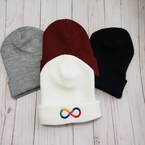 May include: Three beanies in gray, burgundy, white, and black. The white beanie features a rainbow infinity symbol embroidered on the folded brim. The other beanies are solid colors. These knit hats are a comfortable accessory.