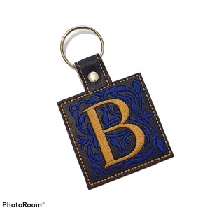 May include: A black leather keychain with a gold-colored letter "B" embroidered on a blue floral design. The keychain has a gold-colored ring and a black strap with a metal rivet.