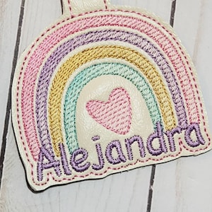 May include: A white felt rainbow with a pink heart in the center and the name "Alejandra" embroidered in purple below.