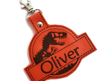 May include: Orange leather keychain with a brown embroidered dinosaur silhouette and the name "Oliver" in brown thread.