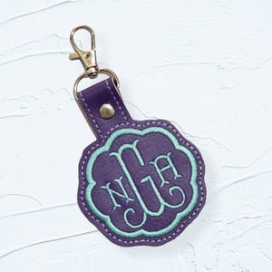 May include: Purple leather keychain with a gold metal clasp and a round, scalloped tag with the monogram "NJA" embroidered in teal.
