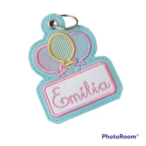 May include: A light blue keychain with a metal ring. The keychain features embroidered balloons in pastel colours and a white rectangle with the name "Emília" in pink script.