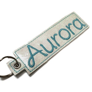 May include: White glitter keychain with a teal blue embroidered name "Aurora" on it.