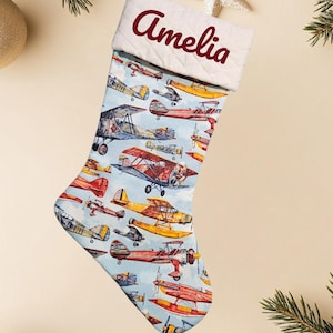 May include: A Christmas stocking with a light blue background featuring various vintage airplanes in red, yellow, and blue. The top of the stocking is white with a quilted pattern and the name "Amelia" in burgundy.