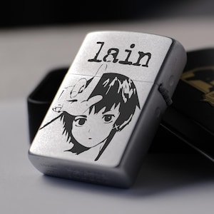 May include: A silver-toned metal lighter featuring a black graphic print of a character with short hair and a hand gesture above their head, with the text "lain" printed above. The lighter is positioned next to its black rectangular storage tin.