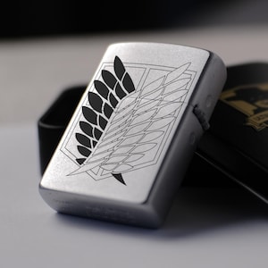 Puede incluir: Un encendedor de metal plateado con un emblema de ala grabado en negro en la parte frontal. El encendedor está colocado junto a su caja de almacenamiento rectangular negra, que muestra el logotipo dorado de Zorro y el nombre de la marca en la tapa.