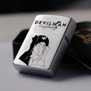 以下が含まれることがあります： 前面に2人のキャラクターの黒いグラフィックと「DEVILMAN crybaby」の文字がプリントされた銀色の金属製ライター。ライターは、ZORROのブランド名が表示された開いた状態の黒い金属製収納ケースの横に置かれています。