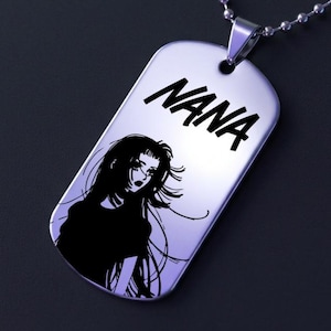 May include: A silver dog tag pendant with the name "NANA" in bold, black letters. Below the name is a black and white illustration of a woman with long hair. The pendant hangs from a silver ball chain.