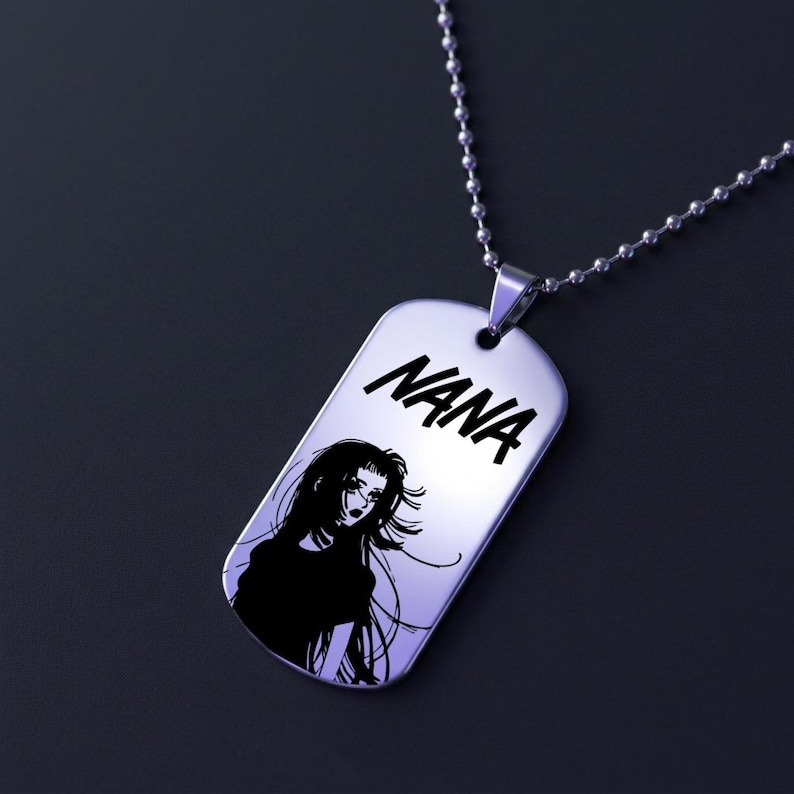 May include: A silver dog tag pendant with the name "NANA" in bold, black letters. Below the name is a black and white illustration of a woman with long hair. The pendant hangs from a silver ball chain.