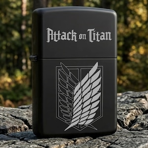 May include: Black lighter with the text "Attack on Titan" and a white emblem of wings. The lighter is rectangular and appears to be made of metal. The background is a blurred view of a forest.