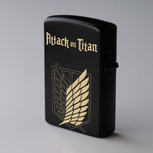 May include: Black lighter with gold text that reads "Attack on Titan" and a gold emblem of wings. The lighter is rectangular and has a hinged lid. The design is a reference to the popular anime series.