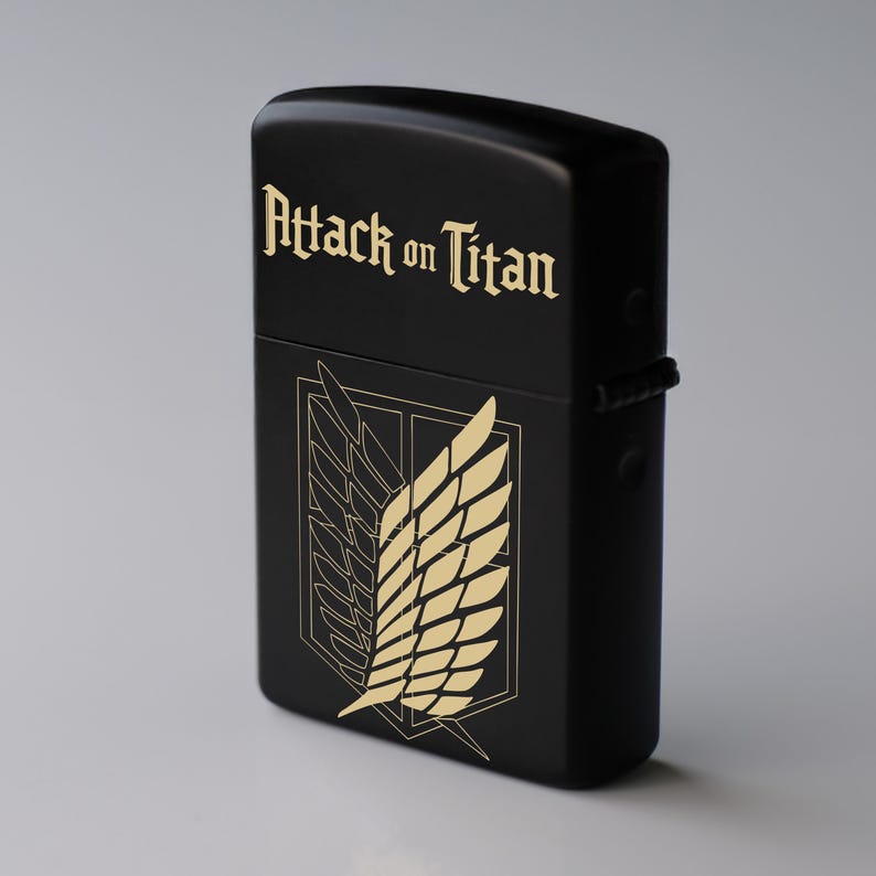 May include: Black lighter with gold text that reads "Attack on Titan" and a gold emblem of wings. The lighter is rectangular and has a hinged lid. The design is a reference to the popular anime series.