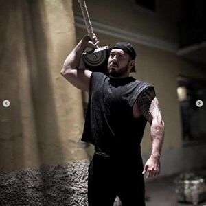 May include: A black sleeveless shirt and black pants worn with a black baseball cap. The outfit is paired with a large silver and black hammer prop featuring a textured handle, held over the shoulder. The ensemble is dark and monochromatic.