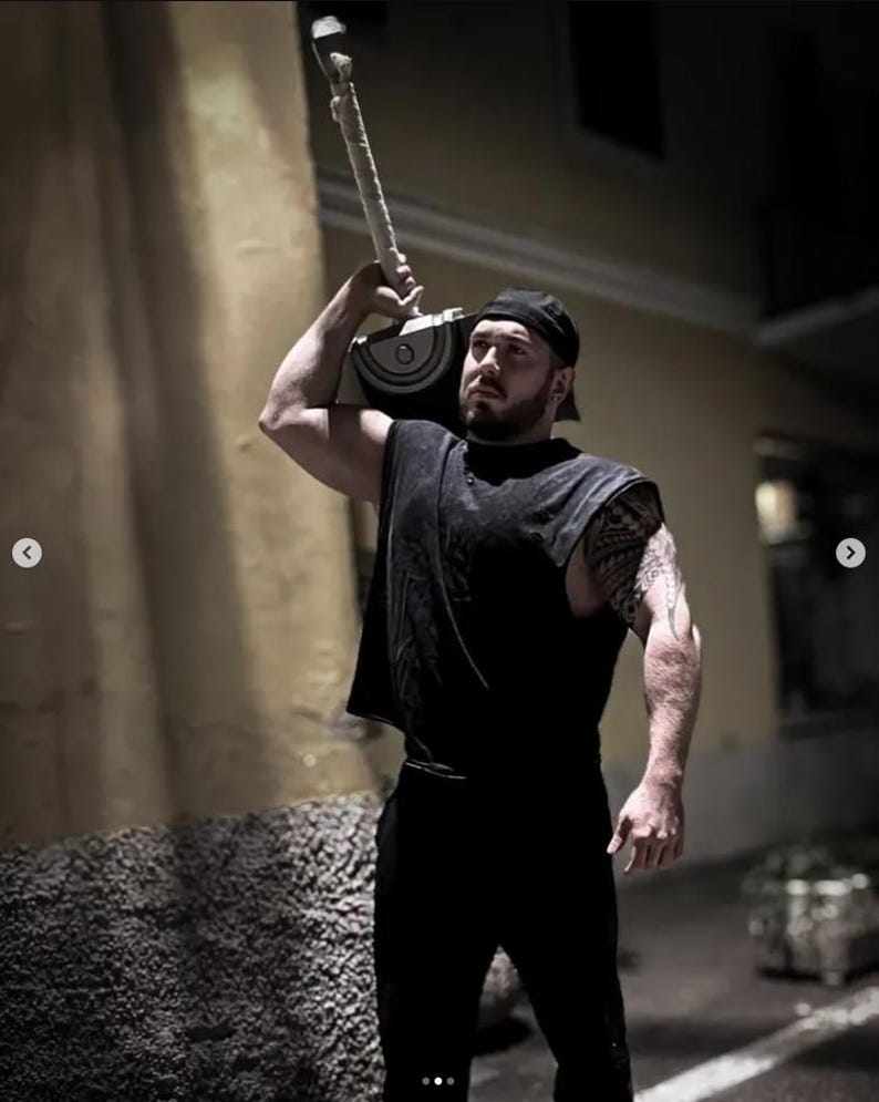 May include: A black sleeveless shirt and black pants worn with a black baseball cap. The outfit is paired with a large silver and black hammer prop featuring a textured handle, held over the shoulder. The ensemble is dark and monochromatic.