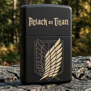 May include: Black lighter with the text "Attack on Titan" in gold lettering. The lighter features a gold emblem of wings on the front. The lighter is sitting on a tree stump.