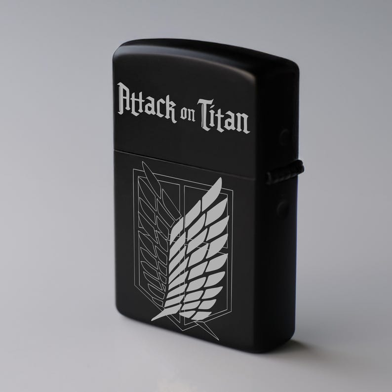 May include: Black lighter with the text "Attack on Titan" in white script. Below the text is a white emblem of wings within a shield. The lighter has a matte finish and a hinged lid.