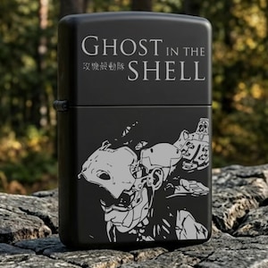 Peut inclure: Briquet noir avec l'inscription blanche "GHOST IN THE SHELL". En dessous, du texte japonais et une illustration grise d'un cyborg. Le briquet est posé sur une roche.