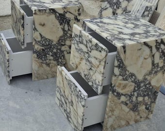 May include: Two marble-topped nightstands with open drawers. The marble has a cream and gray pattern. The drawers are white. The nightstands are cube-shaped with a slightly angled front. The nightstands are on a gray surface.