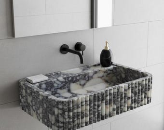 May include: A rectangular wall-mounted bathroom sink crafted from grey and white mottled stone with a fluted exterior texture. The sink features a matte black wall-mounted faucet, a black soap dispenser, and a small white rectangular soap bar.