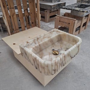 Puede incluir: Un lavabo rectangular de mármol con exterior estriado y desagüe dorado. El lavabo es de color blanquecino con vetas marrones. Está colocado sobre una base de madera, con otros accesorios de mármol al fondo.