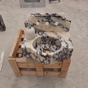 May include: A marble sink with a white and black veined pattern. The sink has a round basin and a decorative backsplash with three tap holes. A matching soap dispenser sits on the side. The sink is displayed on a wooden pallet.
