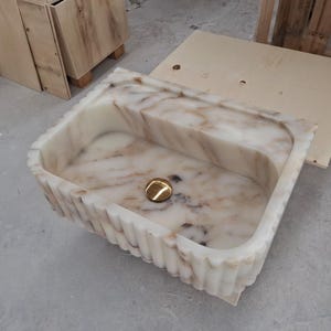 May include: A rectangular farmhouse sink carved from white marble with brown veining. The exterior features a vertical fluted texture, and the interior basin includes a centered, polished gold-toned circular drain cover at the bottom of the basin.