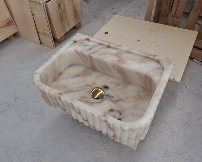 May include: A rectangular farmhouse sink carved from white marble with brown veining. The exterior features a vertical fluted texture, and the interior basin includes a centered, polished gold-toned circular drain cover at the bottom of the basin.