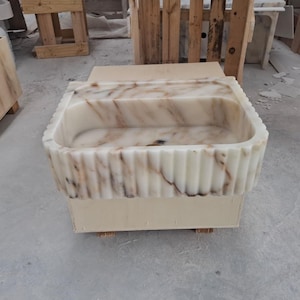 May include: A rectangular stone sink featuring a fluted exterior design with vertical grooves. The basin is crafted from white marble with prominent brown and grey veining throughout the surface, showcasing a polished finish and a deep interior.