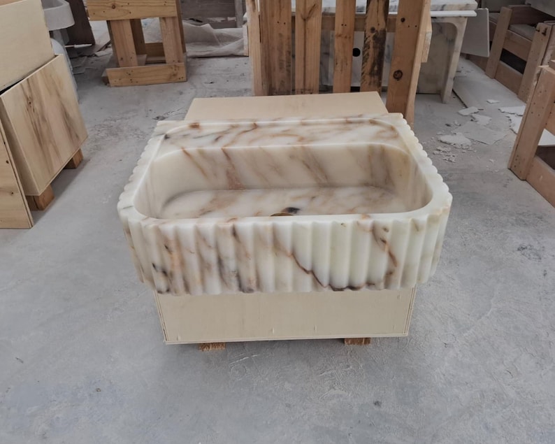 May include: A rectangular stone sink featuring a fluted exterior design with vertical grooves. The basin is crafted from white marble with prominent brown and grey veining throughout the surface, showcasing a polished finish and a deep interior.