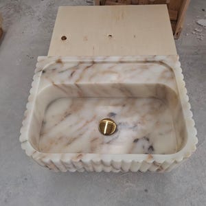 May include: A rectangular marble sink featuring a scalloped, fluted exterior edge and a polished gold-tone drain cover. The stone displays a creamy white base color with prominent, flowing brown and grey veining throughout the basin interior.