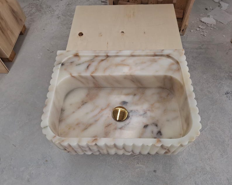 May include: A rectangular marble sink featuring a scalloped, fluted exterior edge and a polished gold-tone drain cover. The stone displays a creamy white base color with prominent, flowing brown and grey veining throughout the basin interior.