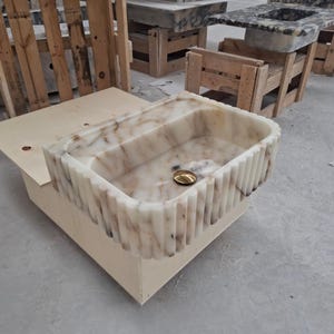 May include: A rectangular stone sink featuring a fluted exterior design with vertical grooves. The basin is crafted from polished white marble with prominent brown veining throughout. A circular gold-toned drain cover is centered in the basin.