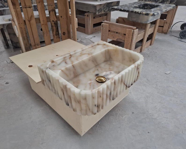 May include: A rectangular stone sink featuring a fluted exterior design with vertical grooves. The basin is crafted from polished white marble with prominent brown veining throughout. A circular gold-toned drain cover is centered in the basin.