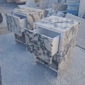 May include: Two marble nightstands with open drawers. The nightstands are white with gray veining, and have a simple, modern design. The drawers are open, revealing the white interior. The nightstands are sitting on a light gray surface.
