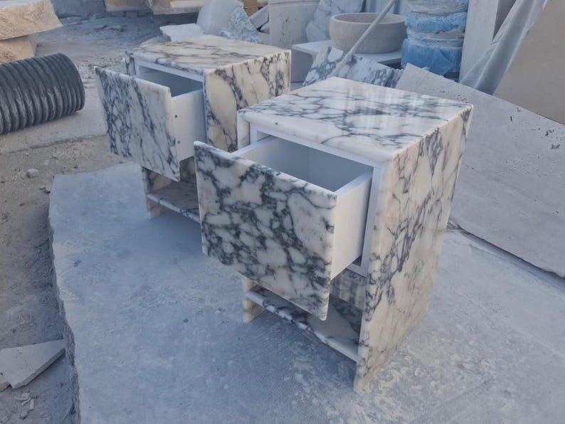 May include: Two marble nightstands with open drawers. The nightstands are white with gray veining, and have a simple, modern design. The drawers are open, revealing the white interior. The nightstands are sitting on a light gray surface.