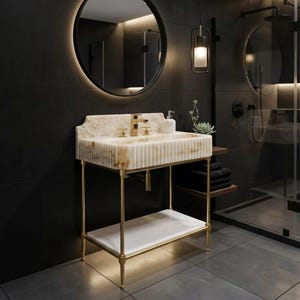 May include: A bathroom vanity featuring a fluted cream-colored stone sink basin with gold-toned metal legs and a lower white shelf. The vanity includes a gold faucet, a matching soap dispenser, and a small succulent plant on the side.
